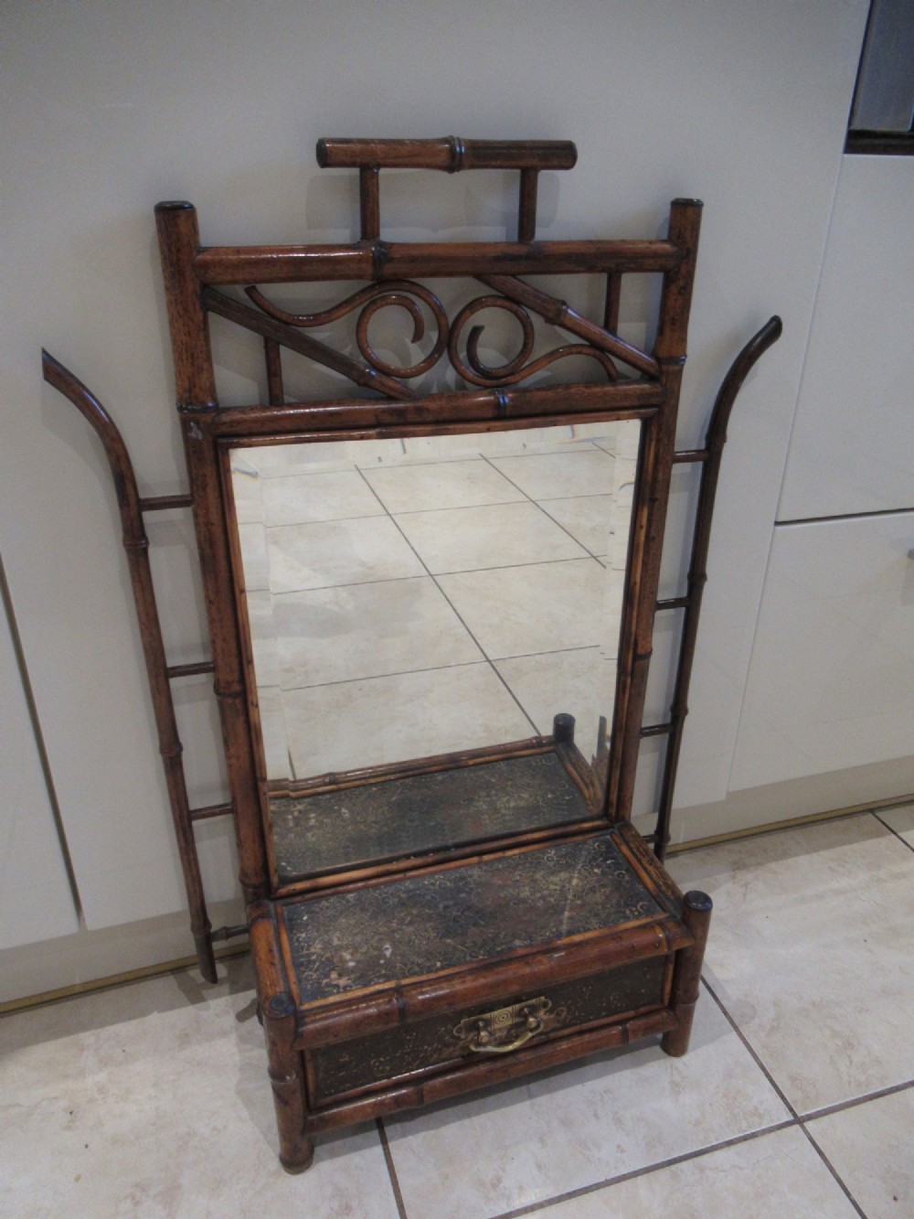 A late Victorian bamboo wall mirror featuring a rectangular bevelled glass plate, bent bamboo scrollwork cresting, side hooks, and a small base drawer with a brass handle. View VII