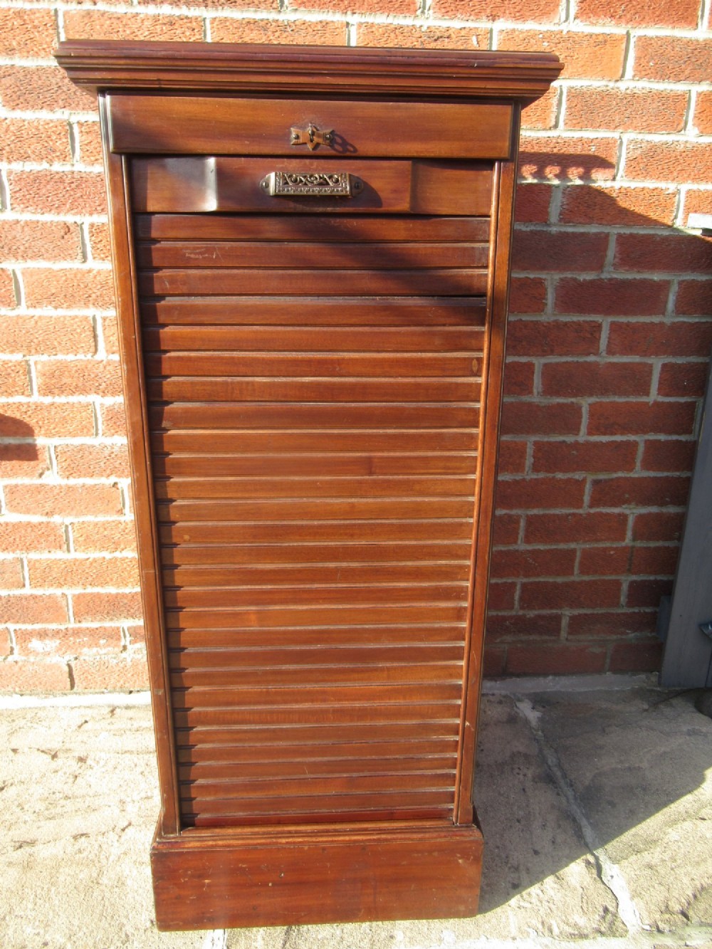 An Edwardian mahogany filing cabinet featuring a slatted tambour roll front door, brass hardware, and a moulded plinth base.
