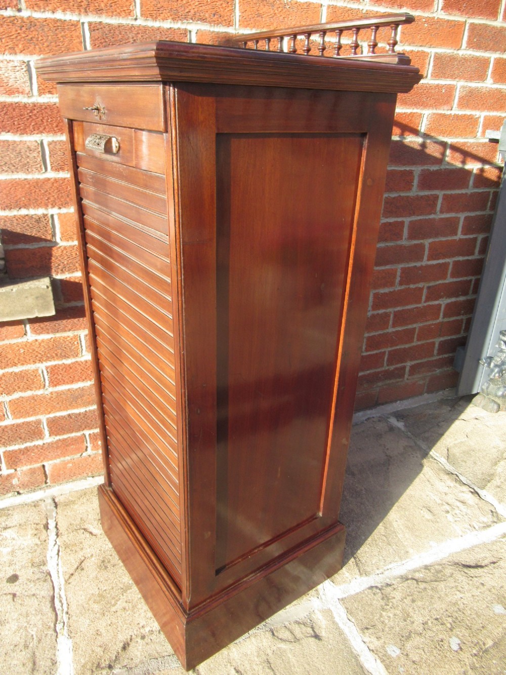 An Edwardian mahogany filing cabinet featuring a slatted tambour roll front door, brass hardware, and a moulded plinth base. View IX