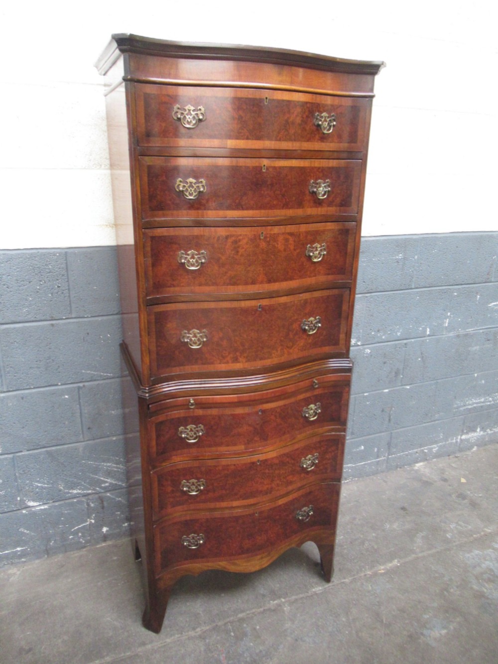 A tall, slim burr walnut chest on chest with a serpentine front, featuring multiple graduated drawers with decorative brass drop handles.