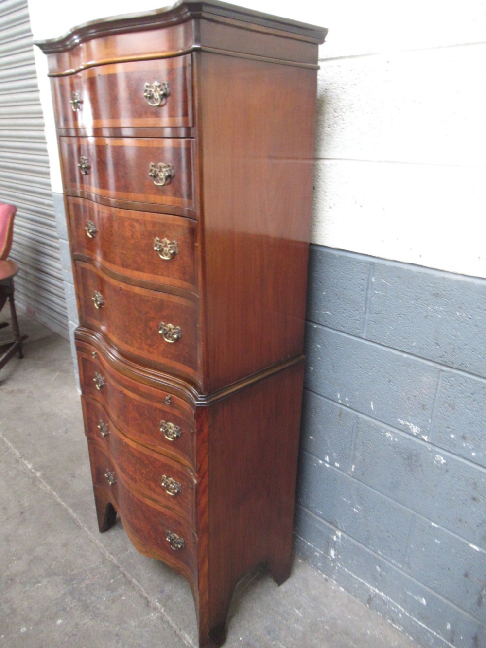 A tall, slim burr walnut chest on chest with a serpentine front, featuring multiple graduated drawers with decorative brass drop handles. View IV