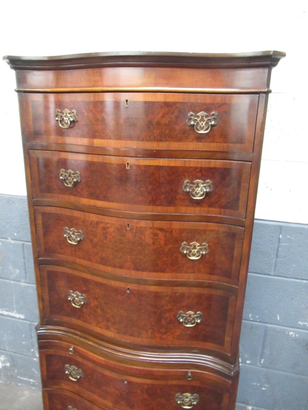 A tall, slim burr walnut chest on chest with a serpentine front, featuring multiple graduated drawers with decorative brass drop handles. View V
