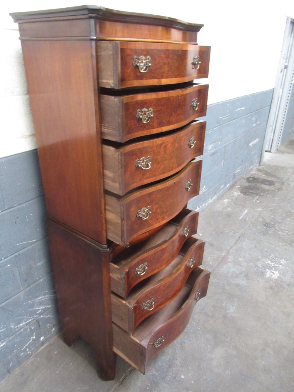 A tall, slim burr walnut chest on chest with a serpentine front, featuring multiple graduated drawers with decorative brass drop handles. View IX