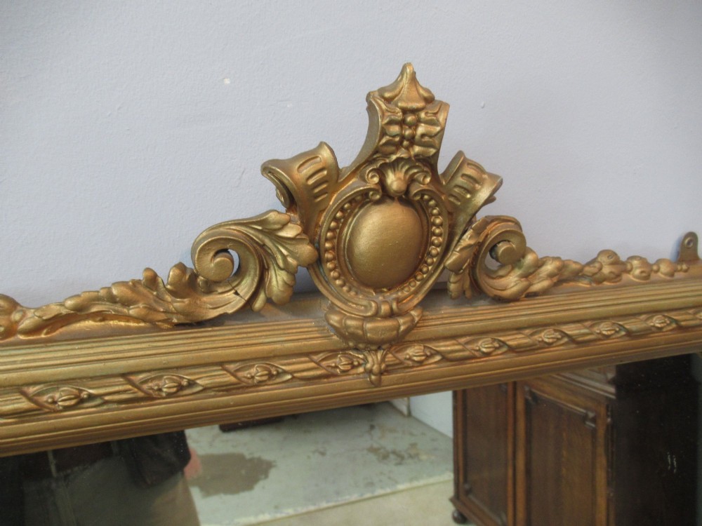 A large rectangular Victorian gilt plaster overmantel mirror featuring an ornate central crest and decorative corner spandrels. View IV