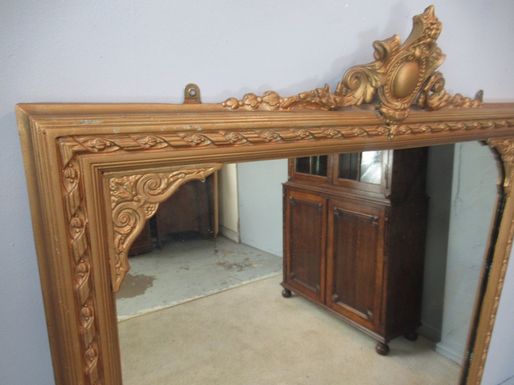 A large rectangular Victorian gilt plaster overmantel mirror featuring an ornate central crest and decorative corner spandrels. View V