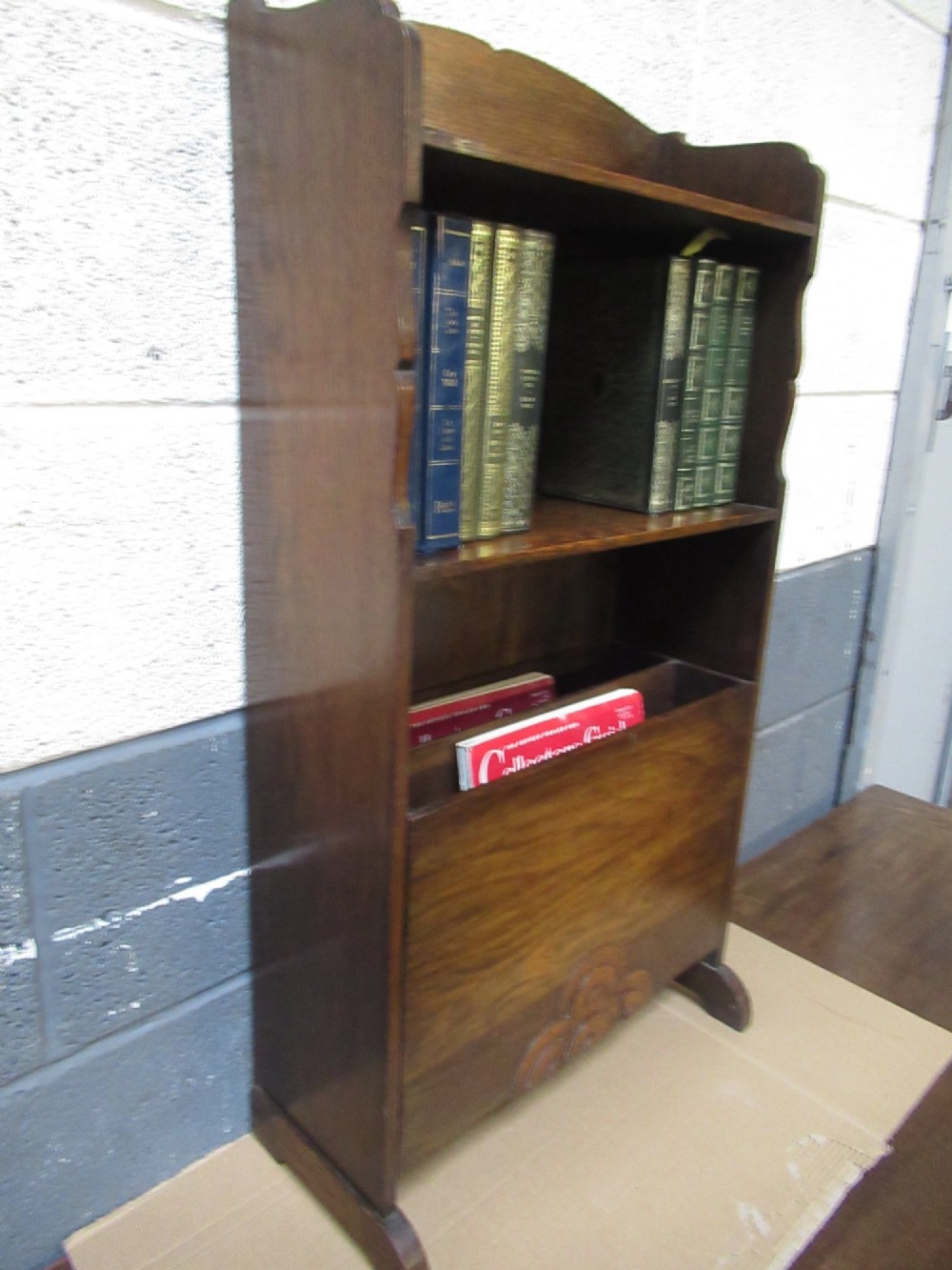 A 1930s dark oak freestanding book rack and magazine holder featuring open shelving above a partitioned lower trough with a carved shell motif. View II
