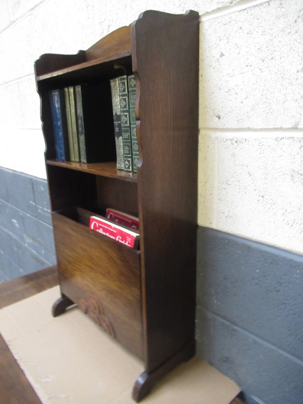 A 1930s dark oak freestanding book rack and magazine holder featuring open shelving above a partitioned lower trough with a carved shell motif. View IX