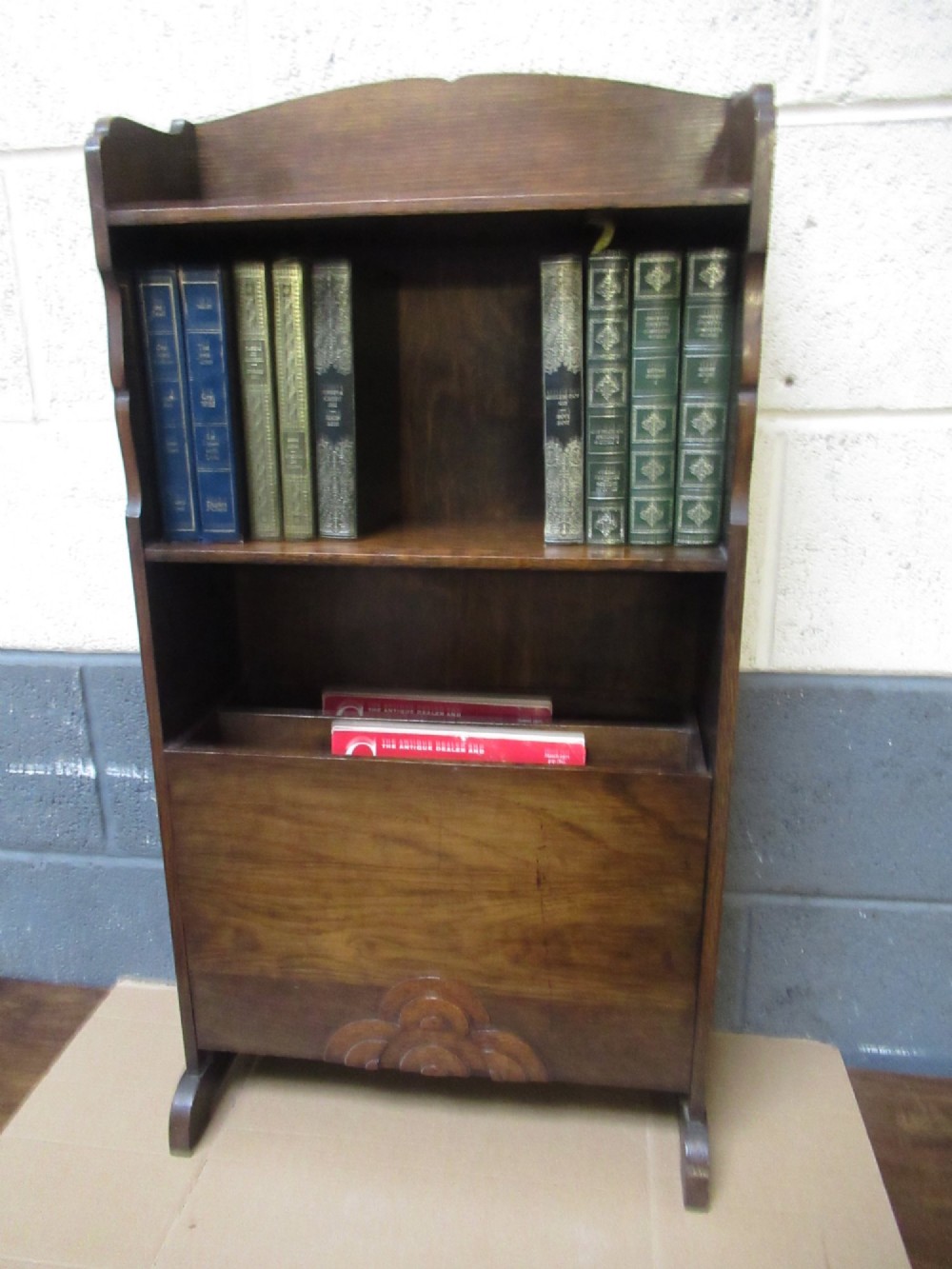 A 1930s dark oak freestanding book rack and magazine holder featuring open shelving above a partitioned lower trough with a carved shell motif. View X