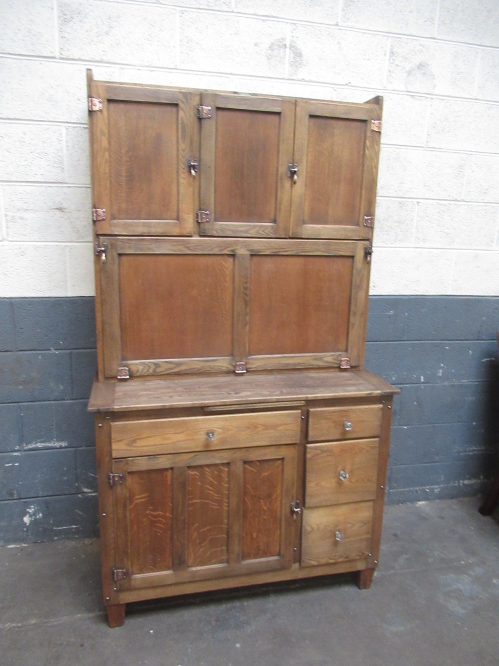 A freestanding 1920s solid oak kitchen cabinet featuring upper and lower panelled cupboards, drawers with glass knobs, and exposed copper hinges. View II