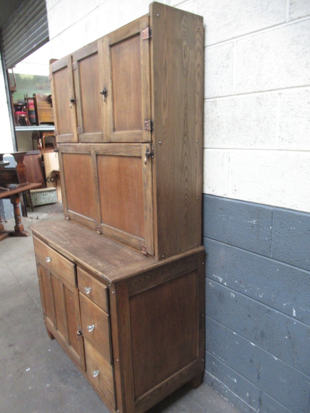 A freestanding 1920s solid oak kitchen cabinet featuring upper and lower panelled cupboards, drawers with glass knobs, and exposed copper hinges. View III