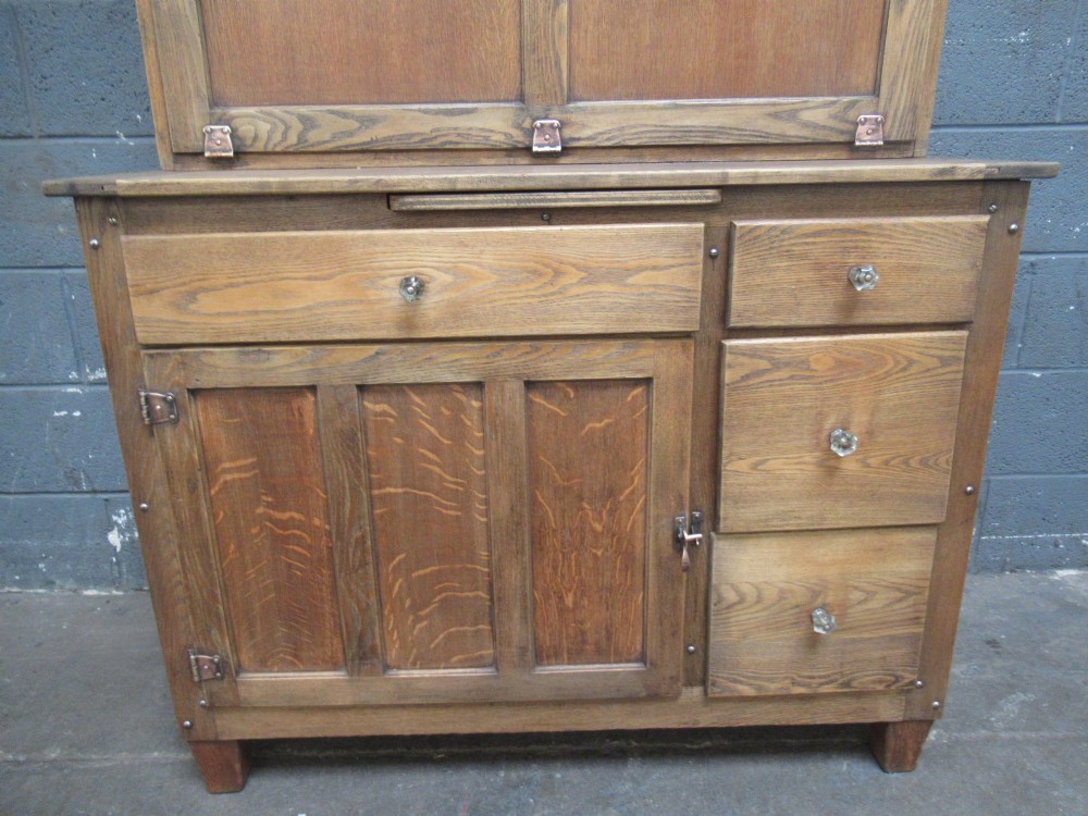 A freestanding 1920s solid oak kitchen cabinet featuring upper and lower panelled cupboards, drawers with glass knobs, and exposed copper hinges. View IV