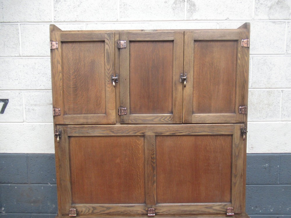 A freestanding 1920s solid oak kitchen cabinet featuring upper and lower panelled cupboards, drawers with glass knobs, and exposed copper hinges. View V