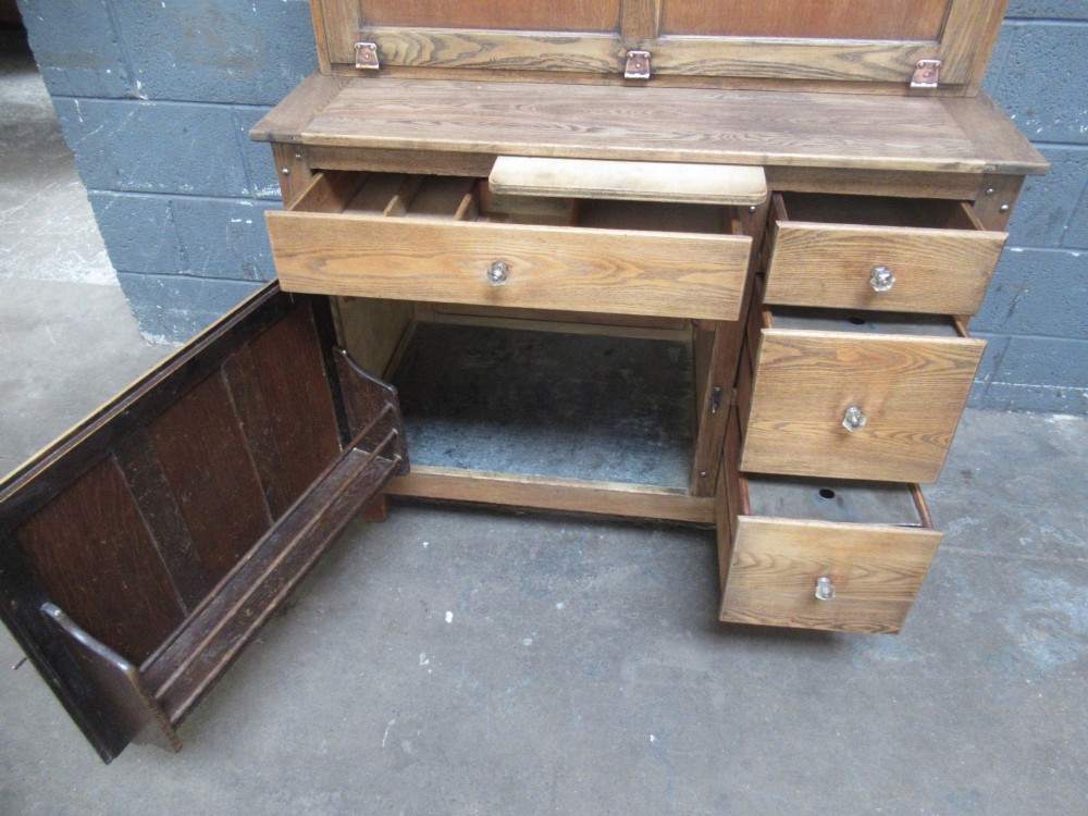 A freestanding 1920s solid oak kitchen cabinet featuring upper and lower panelled cupboards, drawers with glass knobs, and exposed copper hinges. View VI