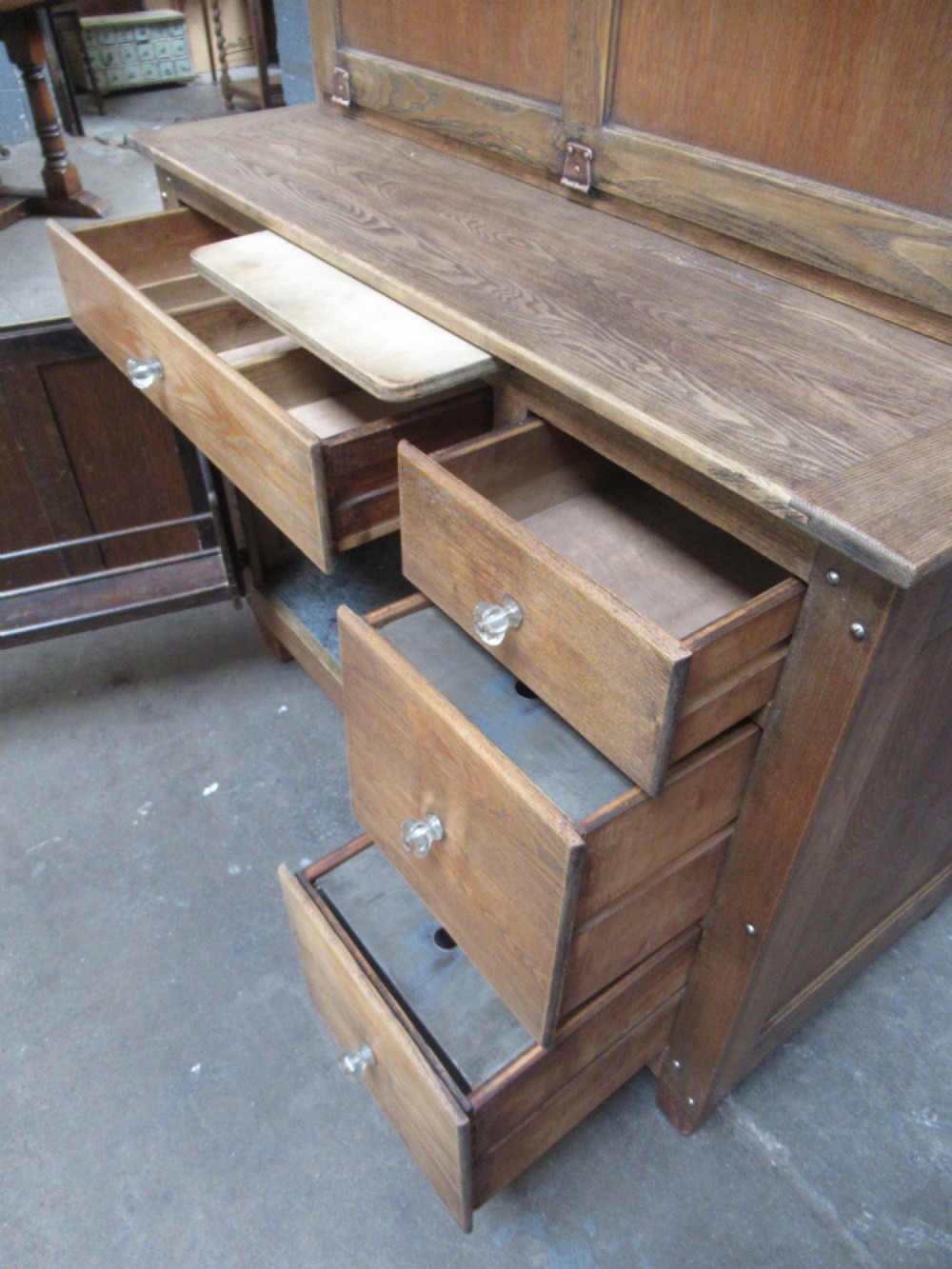 A freestanding 1920s solid oak kitchen cabinet featuring upper and lower panelled cupboards, drawers with glass knobs, and exposed copper hinges. View VIII