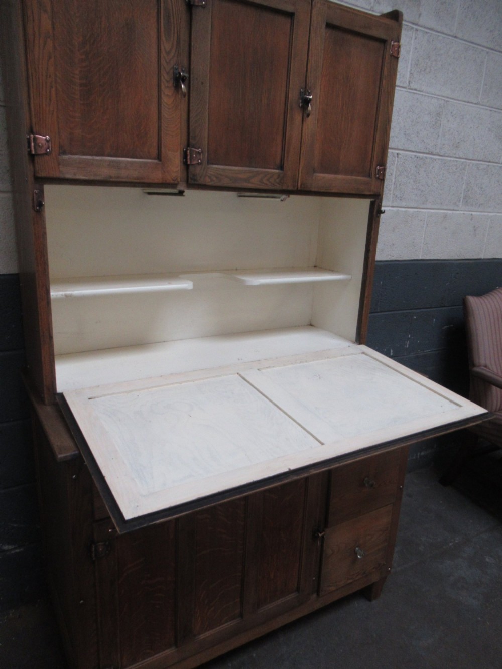 A freestanding 1920s solid oak kitchen cabinet featuring upper and lower panelled cupboards, drawers with glass knobs, and exposed copper hinges. View IX