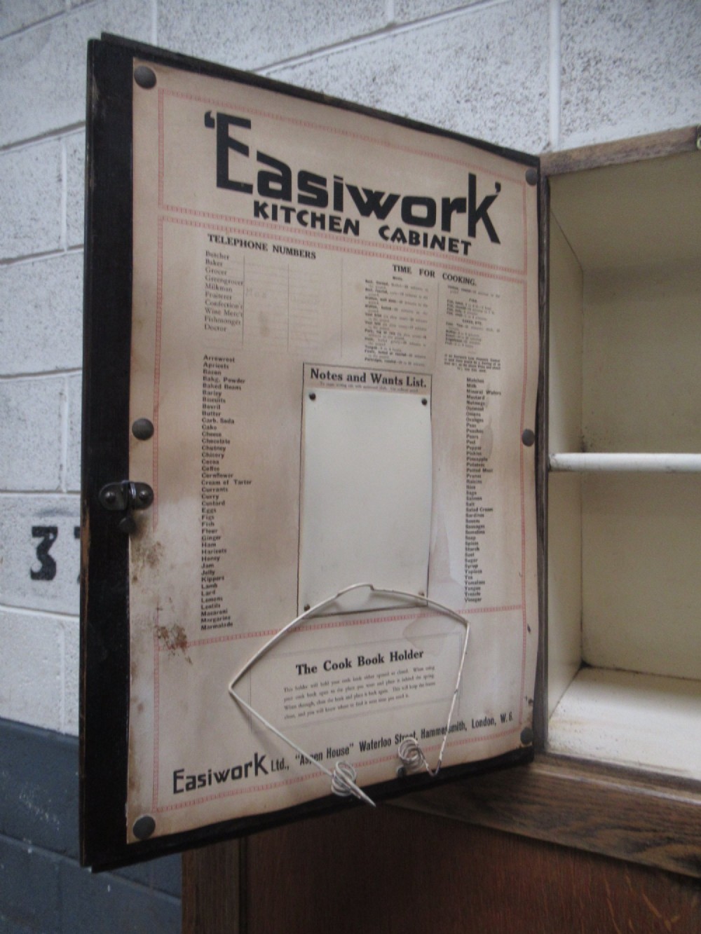 A freestanding 1920s solid oak kitchen cabinet featuring upper and lower panelled cupboards, drawers with glass knobs, and exposed copper hinges. View XII
