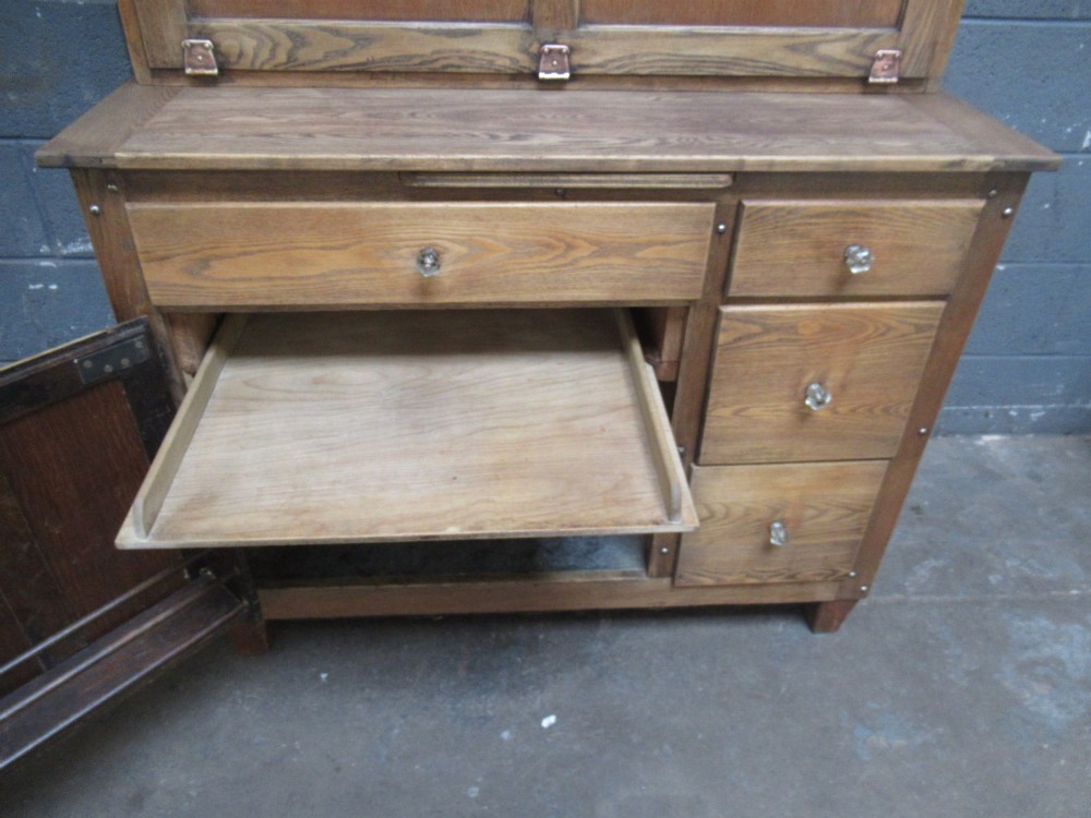 A freestanding 1920s solid oak kitchen cabinet featuring upper and lower panelled cupboards, drawers with glass knobs, and exposed copper hinges. View XIV