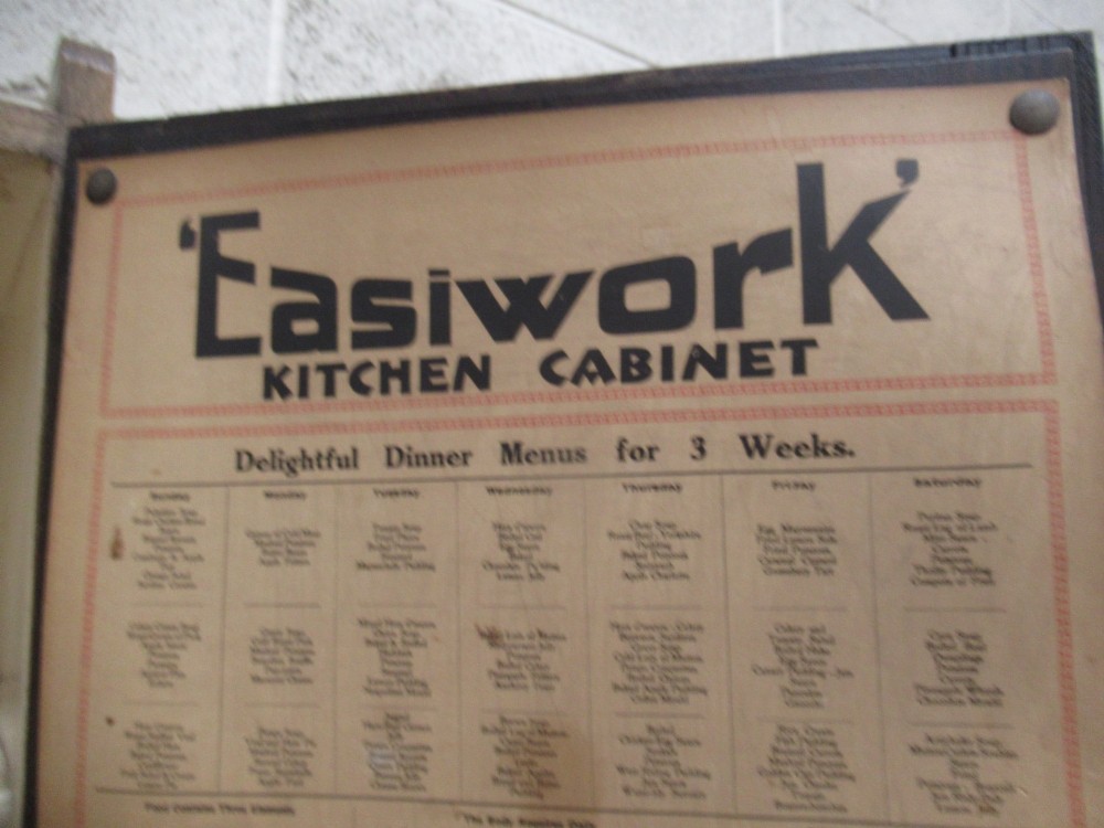 A freestanding 1920s solid oak kitchen cabinet featuring upper and lower panelled cupboards, drawers with glass knobs, and exposed copper hinges. View XV