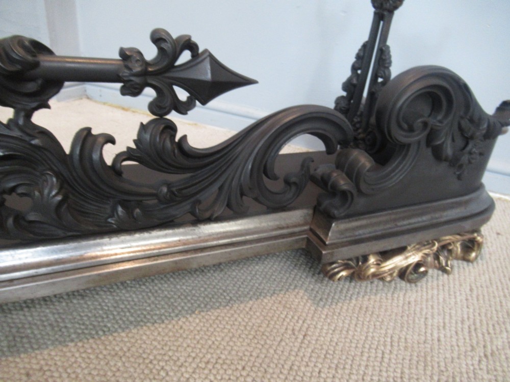 A nineteenth-century cast iron fire fender featuring detailed scrolling foliage ornament, a central spear-head rail, and brass feet. View VI