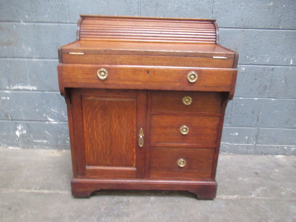 A compact warm brown oak roll top desk featuring a slatted tambour top section, brass ring drawer knobs, a panelled left cupboard, and three stacked side drawers.