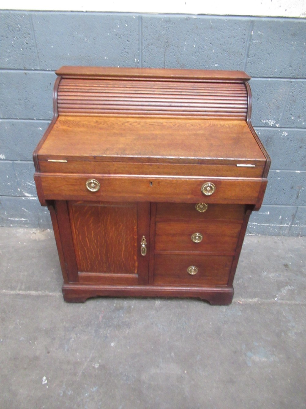 A compact warm brown oak roll top desk featuring a slatted tambour top section, brass ring drawer knobs, a panelled left cupboard, and three stacked side drawers. View II