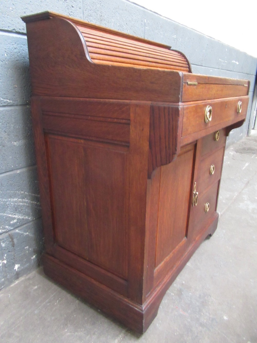 A compact warm brown oak roll top desk featuring a slatted tambour top section, brass ring drawer knobs, a panelled left cupboard, and three stacked side drawers. View III