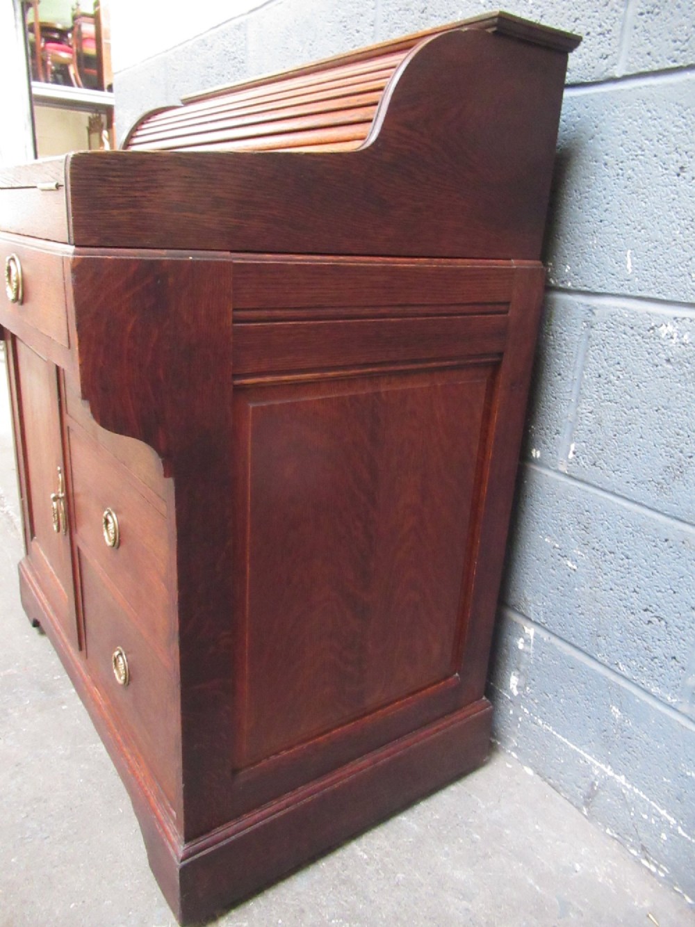 A compact warm brown oak roll top desk featuring a slatted tambour top section, brass ring drawer knobs, a panelled left cupboard, and three stacked side drawers. View IV