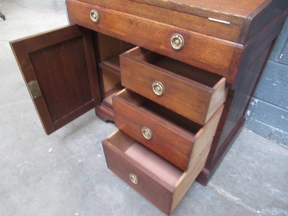 A compact warm brown oak roll top desk featuring a slatted tambour top section, brass ring drawer knobs, a panelled left cupboard, and three stacked side drawers. View VI