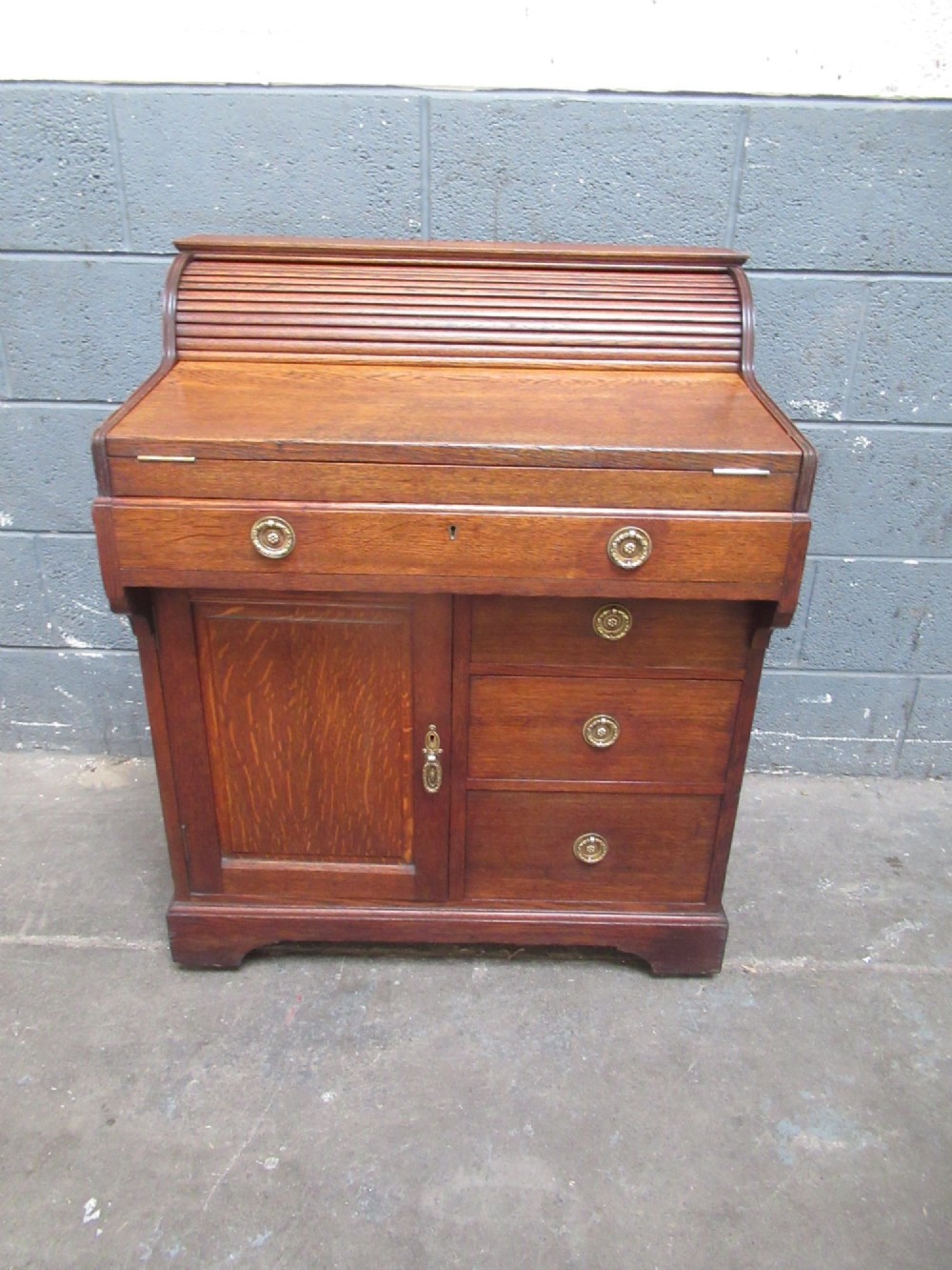 A compact warm brown oak roll top desk featuring a slatted tambour top section, brass ring drawer knobs, a panelled left cupboard, and three stacked side drawers. View XIV