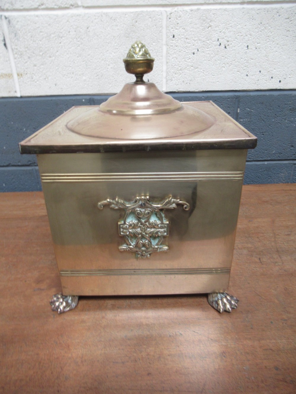 A square brass coal bin featuring a domed copper-toned lid topped with a finial, side drop ring handles, a relief crest, and claw feet. View II