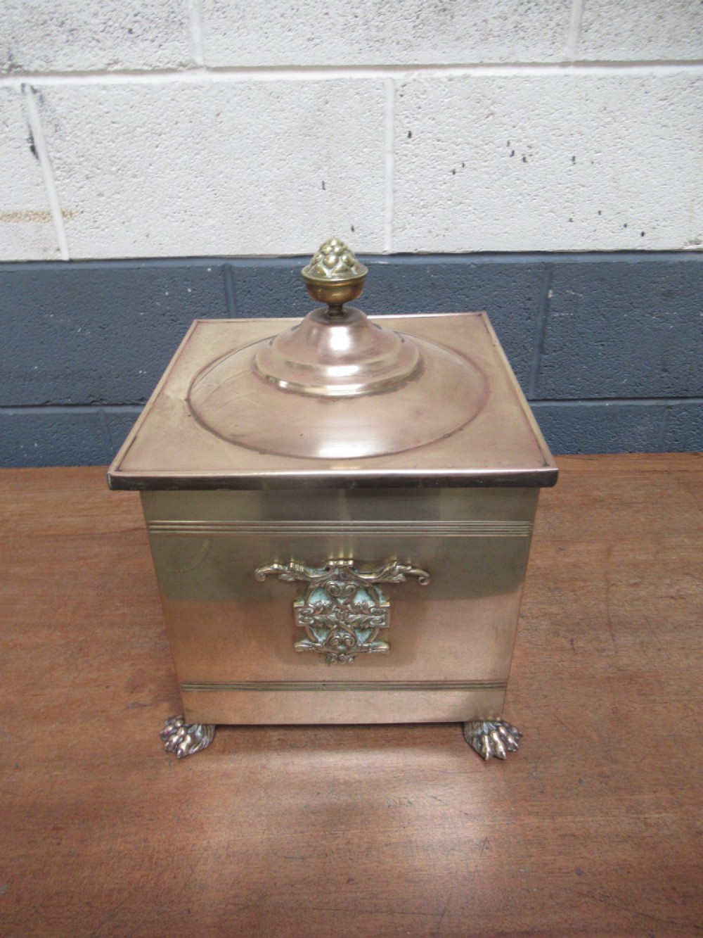 A square brass coal bin featuring a domed copper-toned lid topped with a finial, side drop ring handles, a relief crest, and claw feet. View III