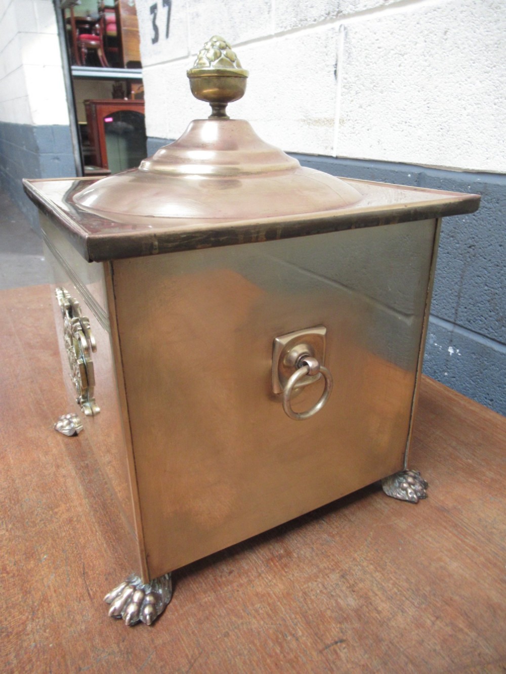 A square brass coal bin featuring a domed copper-toned lid topped with a finial, side drop ring handles, a relief crest, and claw feet. View IV