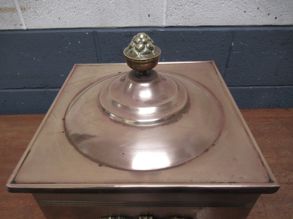 A square brass coal bin featuring a domed copper-toned lid topped with a finial, side drop ring handles, a relief crest, and claw feet. View V