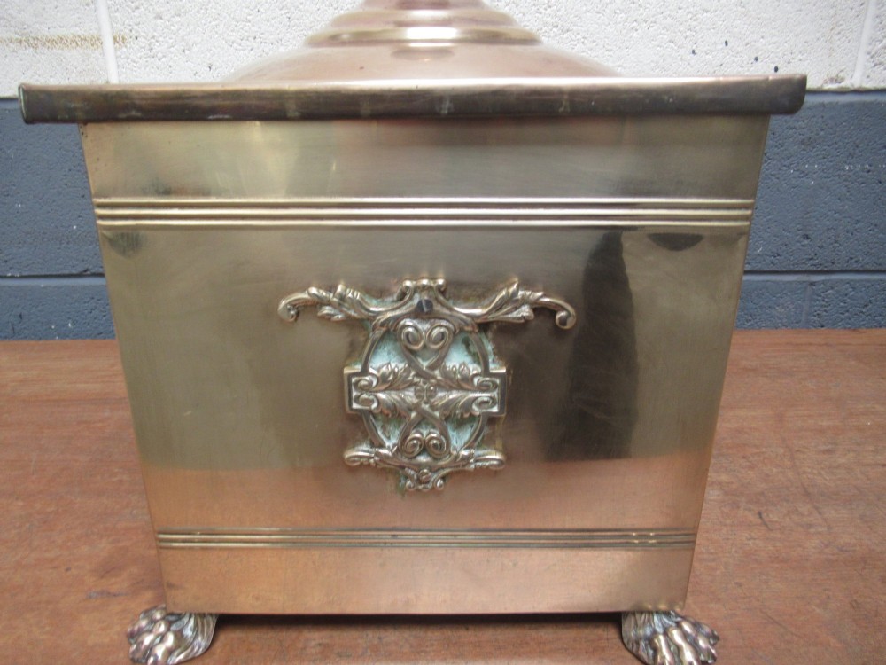 A square brass coal bin featuring a domed copper-toned lid topped with a finial, side drop ring handles, a relief crest, and claw feet. View VI