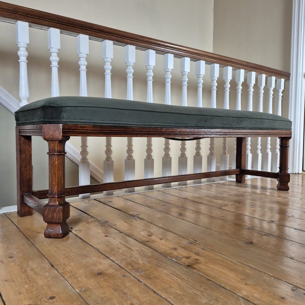 A long Victorian Gothic Revival solid oak bench featuring turned legs, a rectangular lower stretcher frame, and a dark green velvet upholstered cushion top. View V
