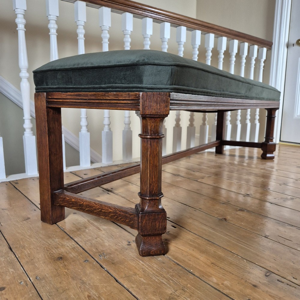 A long Victorian Gothic Revival solid oak bench featuring turned legs, a rectangular lower stretcher frame, and a dark green velvet upholstered cushion top. View VI