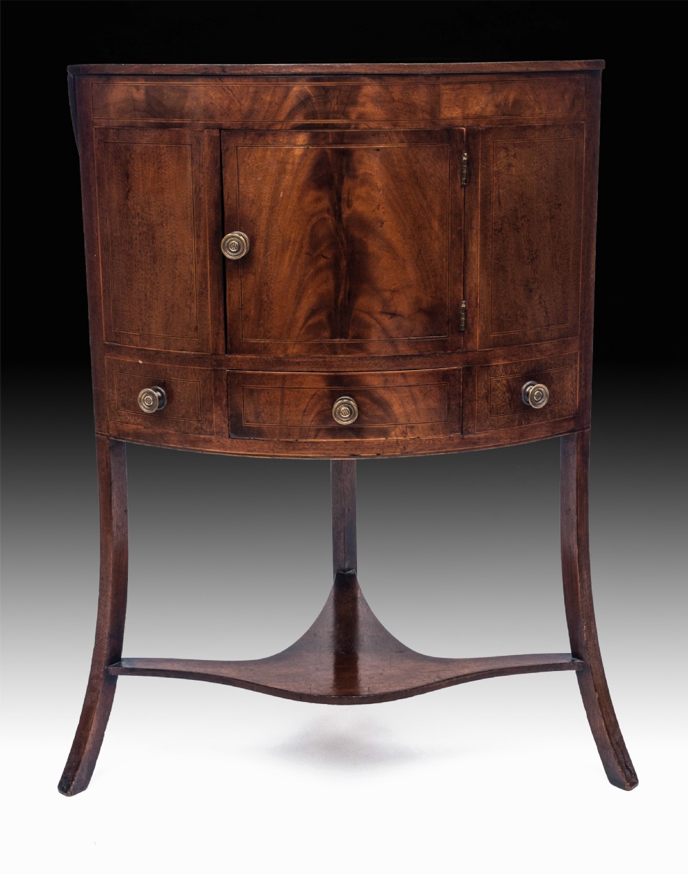 A Georgian mahogany corner washstand featuring a curved central door, brass knob handles, inlaid boxwood stringing, and splayed legs connected by a shaped undershelf.