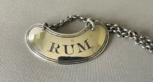 fig 1. A kidney-shaped silver decanter label engraved with the word RUM and attached to a silver link chain.