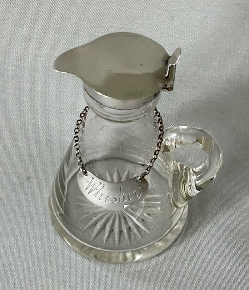 fig 1. A clear glass conical whiskey tot featuring a hinged silver lid, a side handle, a star-cut base, and a silver crescent label engraved with Whiskey attached by a chain around the neck.