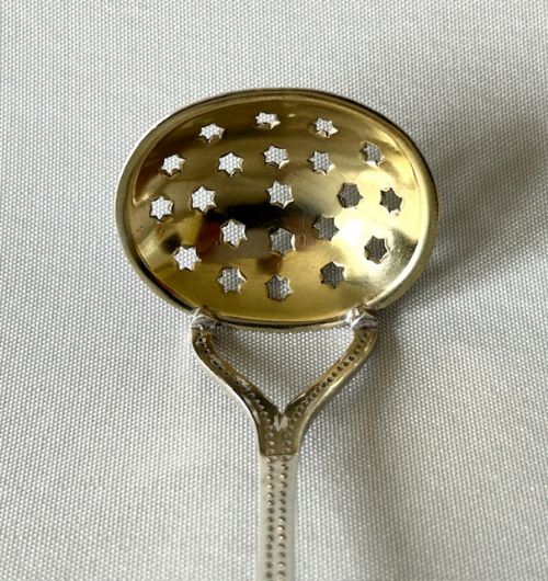 fig 1. An Arts and Crafts silver gilt sifter spoon featuring star-shaped perforations in an oval bowl and a beaded edge handle.