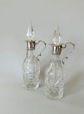 fig 1. A pair of Victorian cut glass oil and vinegar bottles featuring sterling silver collars, scroll handles, and faceted glass stoppers.
