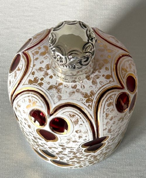 An Edwardian Bohemian glass scent bottle featuring ruby red cut-outs over a white gilded pattern, topped with a hallmarked silver cap. View VI