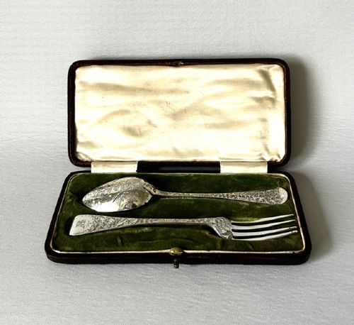 A late Victorian silver spoon and fork with engraved pattern detailing, displayed inside an open dark green velvet-lined presentation box with a cream silk interior.
