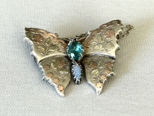 A Victorian sterling silver butterfly brooch detailed with applied rose gold stars and floral motifs, set with a central oval green stone. View V