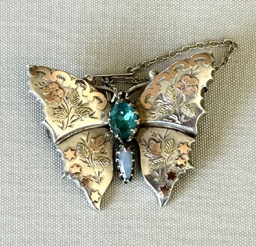 A Victorian sterling silver butterfly brooch detailed with applied rose gold stars and floral motifs, set with a central oval green stone. View VI