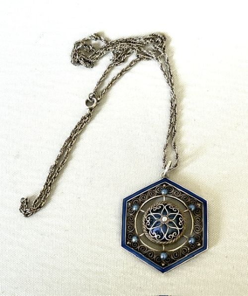 fig 1. A hexagonal silver pendant with delicate wire filigree scrolls surrounding a central blue guilloche enamel dome, suspended from a silver chain.