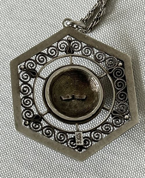 fig 3. A hexagonal silver pendant with delicate wire filigree scrolls surrounding a central blue guilloche enamel dome, suspended from a silver chain.