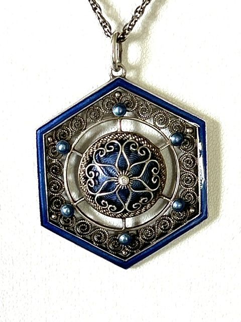 fig 4. A hexagonal silver pendant with delicate wire filigree scrolls surrounding a central blue guilloche enamel dome, suspended from a silver chain.