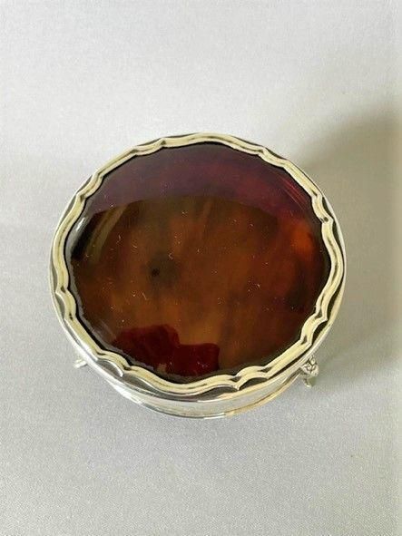 fig 1. A circular George V sterling silver trinket box featuring a dark tortoiseshell lid with a scalloped edge resting on three silver legs.