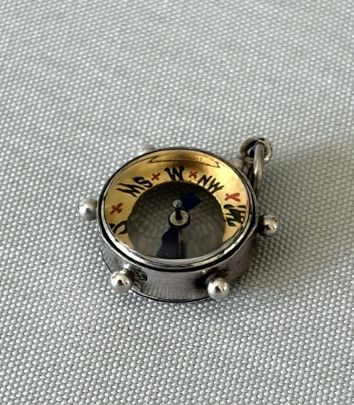 fig 1. A miniature silver fob compass designed in the shape of a ships wheel with a central directional needle and suspension ring.